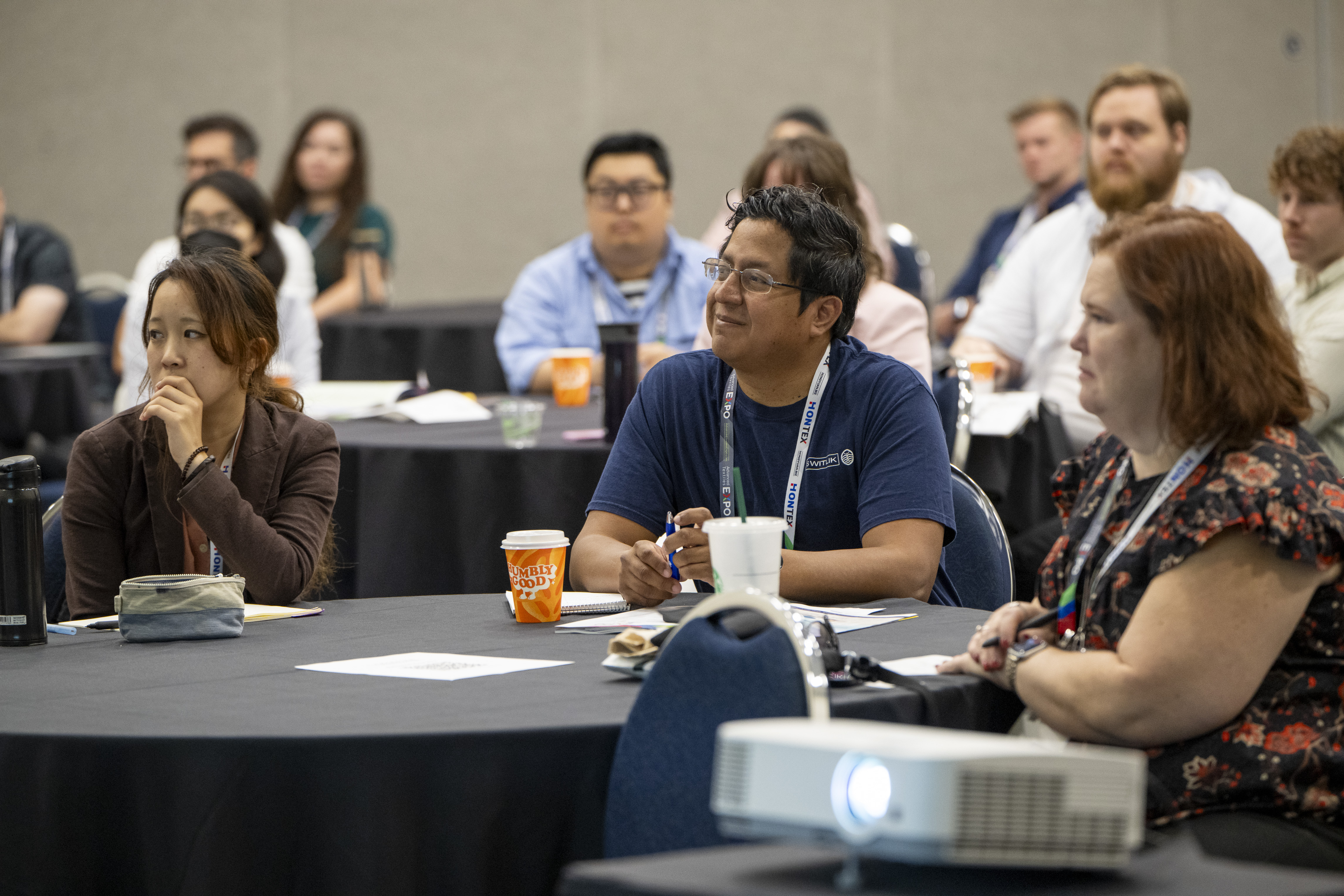 Attendees at a conference sit at tables, attentively listening. The room features dark tables with papers, drinks, and a projector.