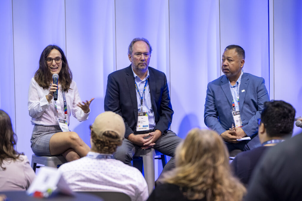 Panel discussion featuring three speakers: a woman with glasses actively gesturing, a man in a suit listening, and another in a blue blazer.