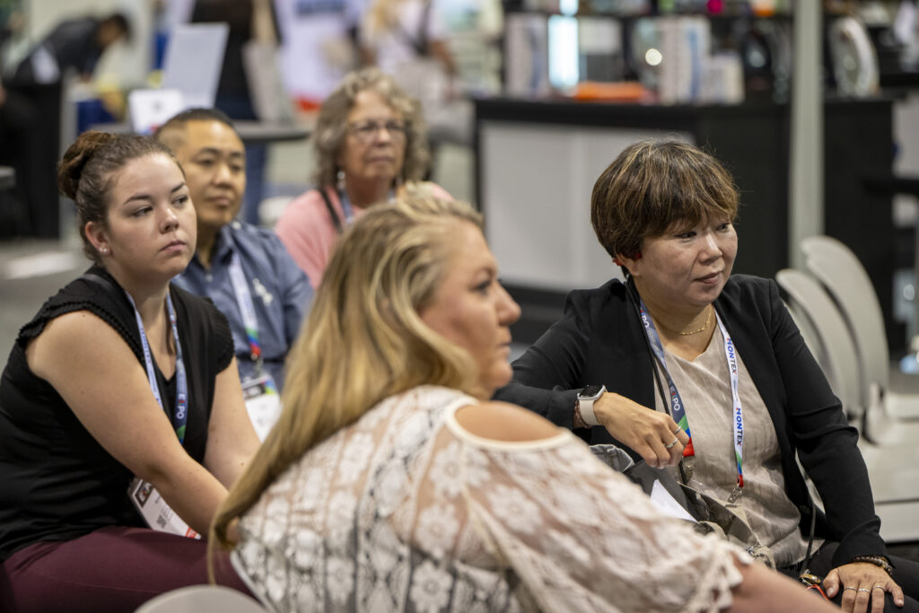 A group of five attendees sitting closely, engaged in discussion; varied hairstyles and outfits, with a colorful conference backdrop.