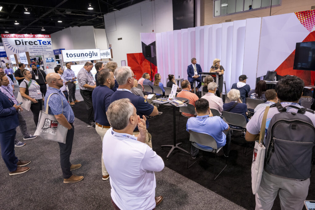 A crowd engages in a presentation at a conference, with a speaker at a podium and seated panelists against a modern backdrop.
