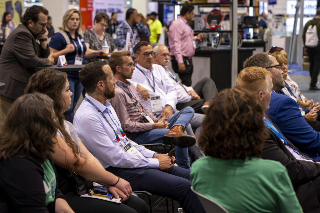Attendees engaged in discussion at a conference, diverse group seated in rows, wearing badges, with event displays in the background.
