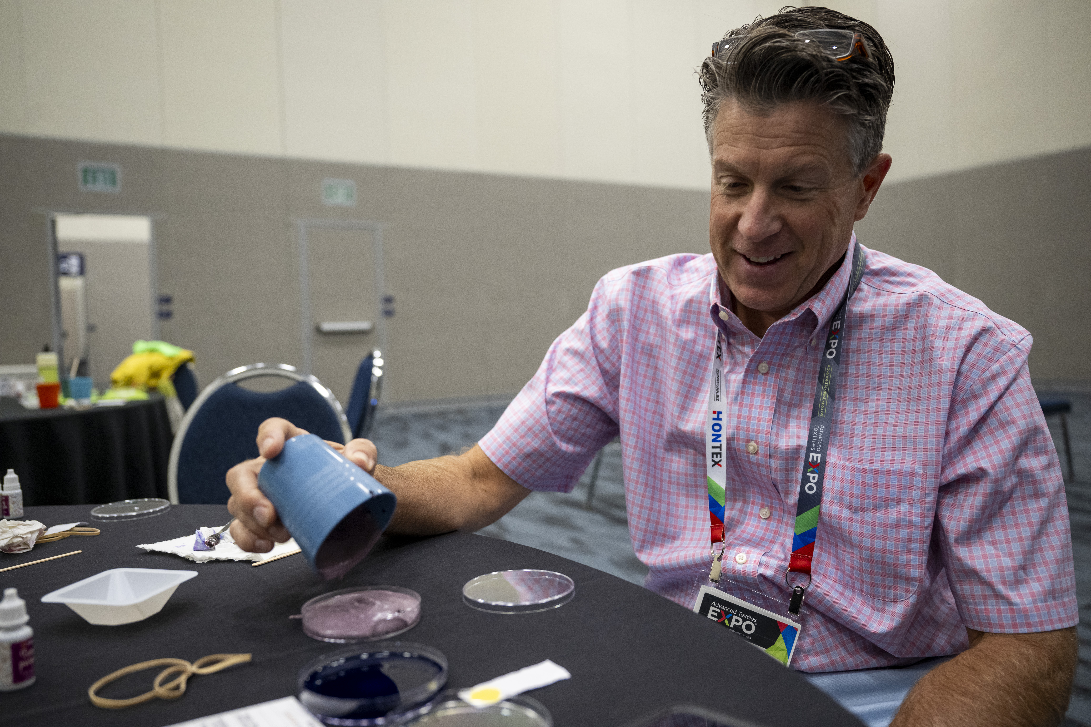 A man in a pink checkered shirt tilts a blue cup over a table, examining purple mixture, surrounded by various workshop materials.