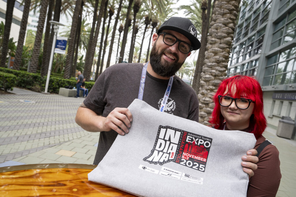Two smiling individuals hold a grey tote bag featuring the Advanced Textiles Expo 2025 logo, set against palm trees and a modern building.