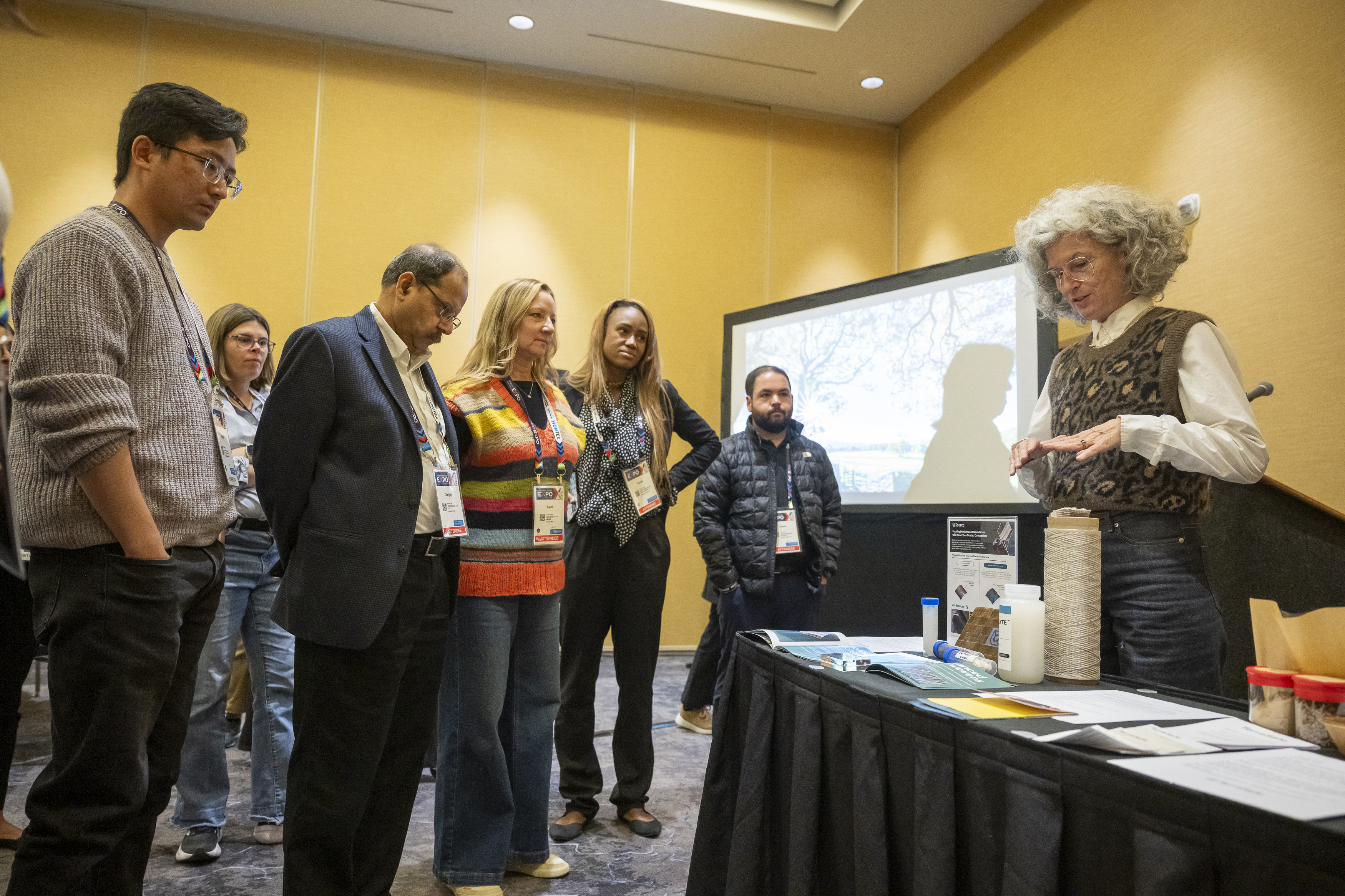 Participants gather around a table displaying textile samples and materials, while a presenter discusses innovations in a modern conference room.
