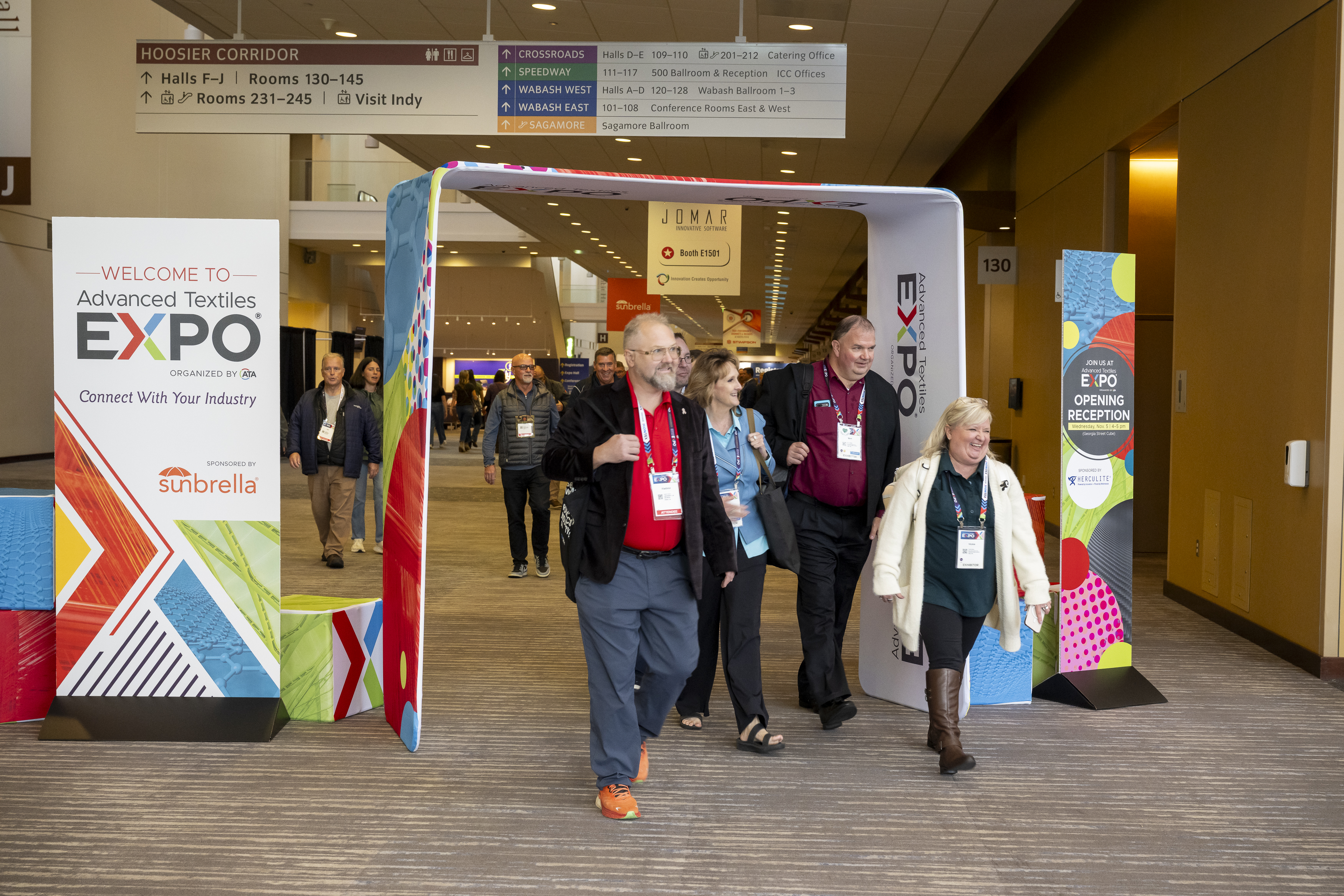 Five attendees walk through a colorful archway welcoming visitors to the Advanced Textiles Expo, with directional signs above and vibrant banners on the sides.