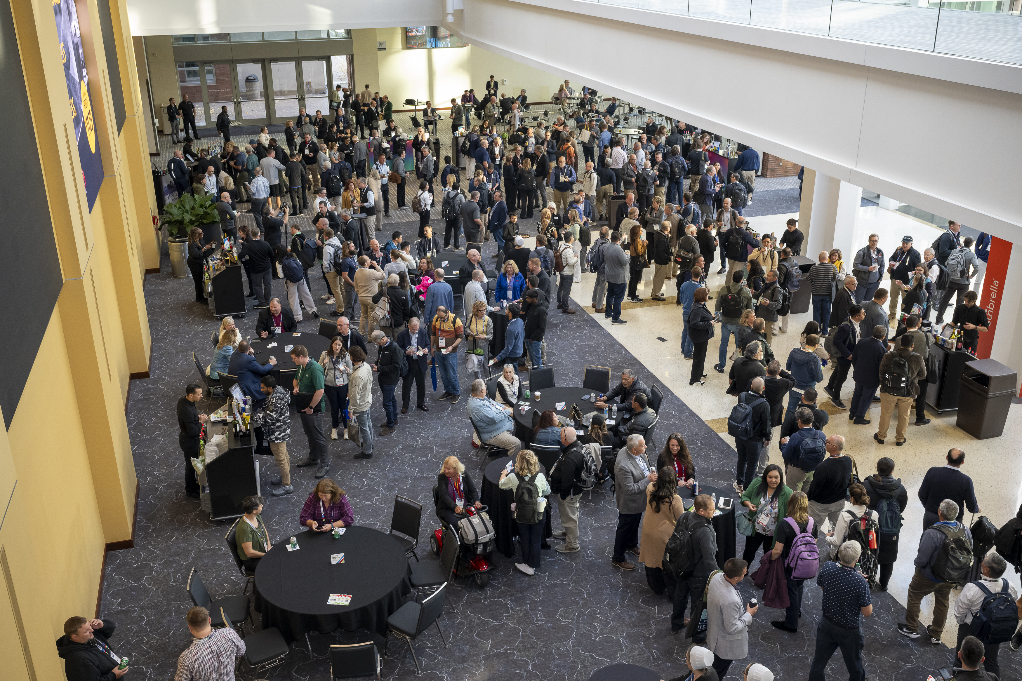 Crowded conference hall filled with attendees engaging around round tables, with a backdrop of large windows and banners.