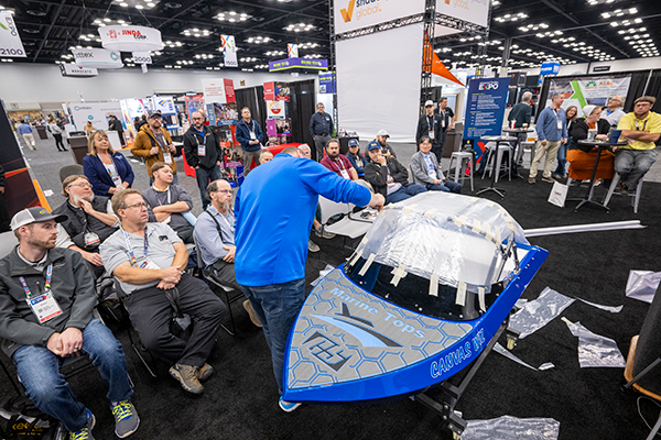 Demonstration of a sleek blue boat model by a presenter in a blue sweater, with an engaged audience observing in a trade show setting.