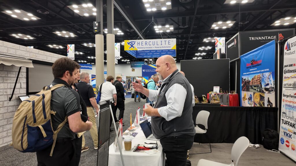 A trade show scene featuring a vendor in a gray vest speaking with attendees, colorful banners overhead, and various displays in the background.