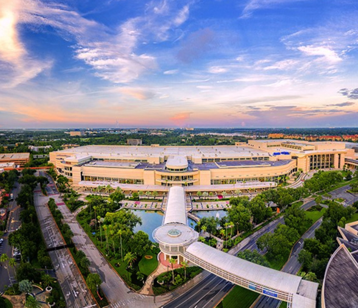 Sprawling modern building surrounded by greenery, with a curved walkway crossing a pond, under a vibrant sky at dusk.