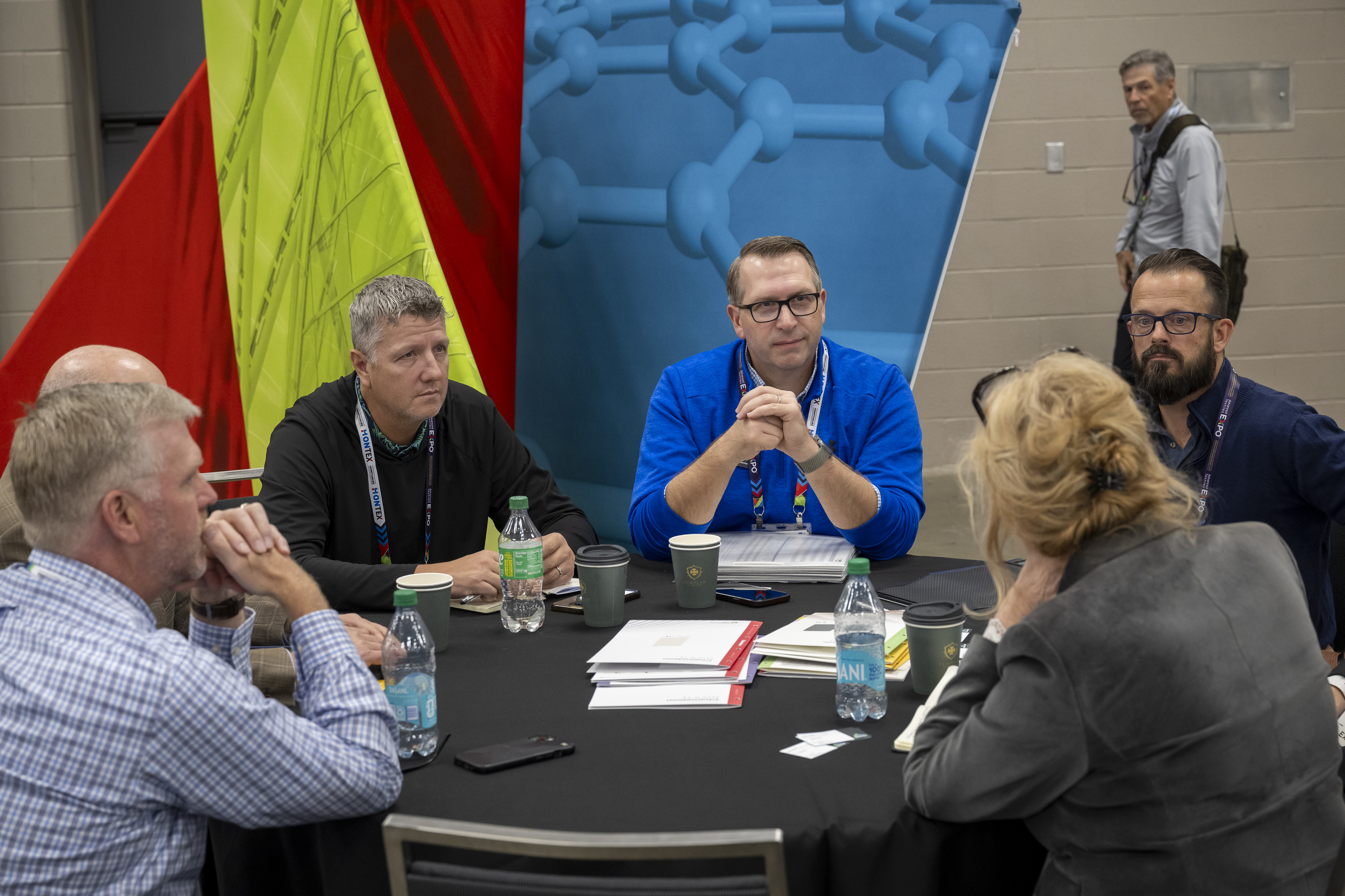 Participants engaged in conversation around a black table, surrounded by colorful backdrop displays. Papers and drinks are present.