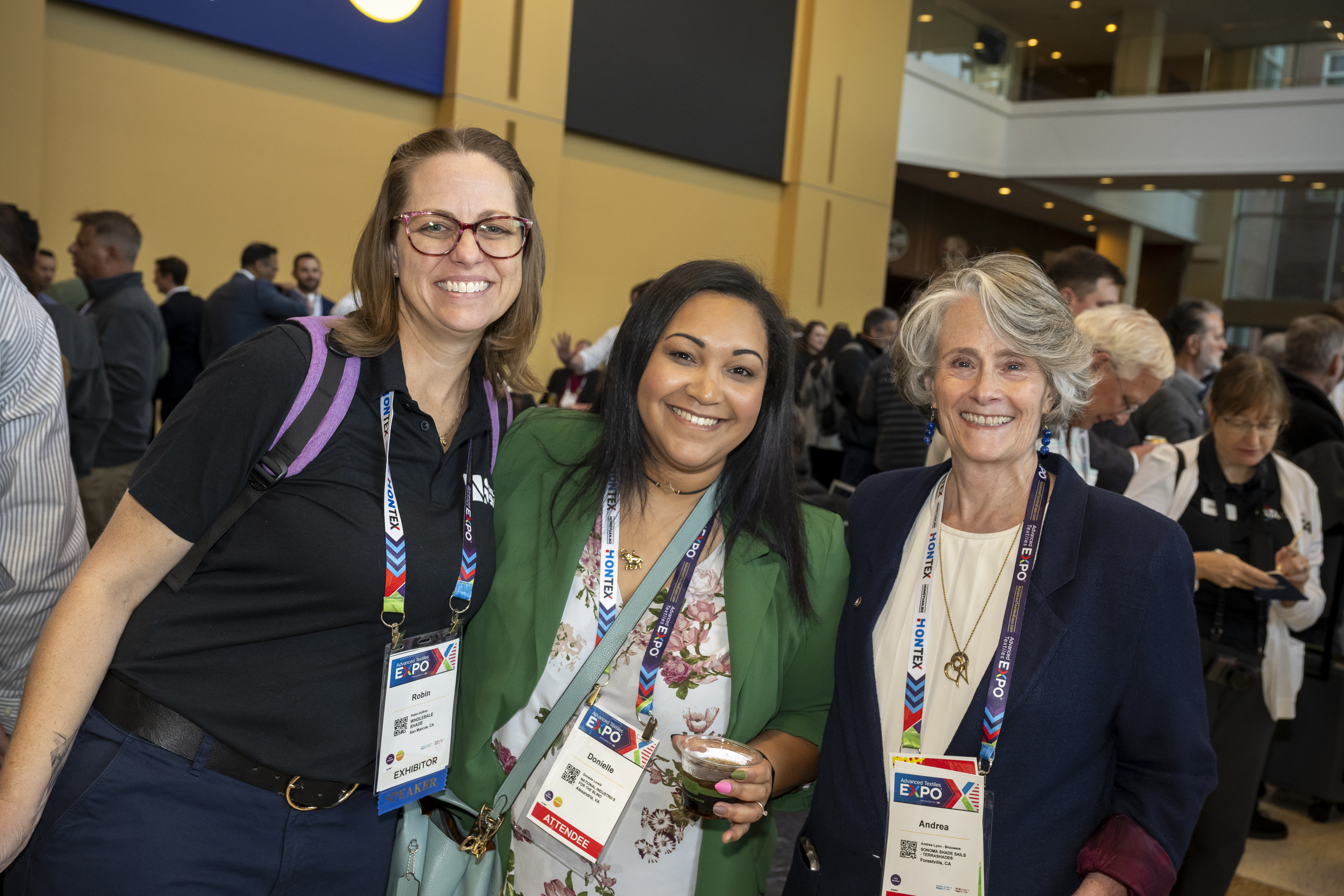 Three women smile together at a conference, wearing badges, with a lively background of attendees in a spacious indoor venue.