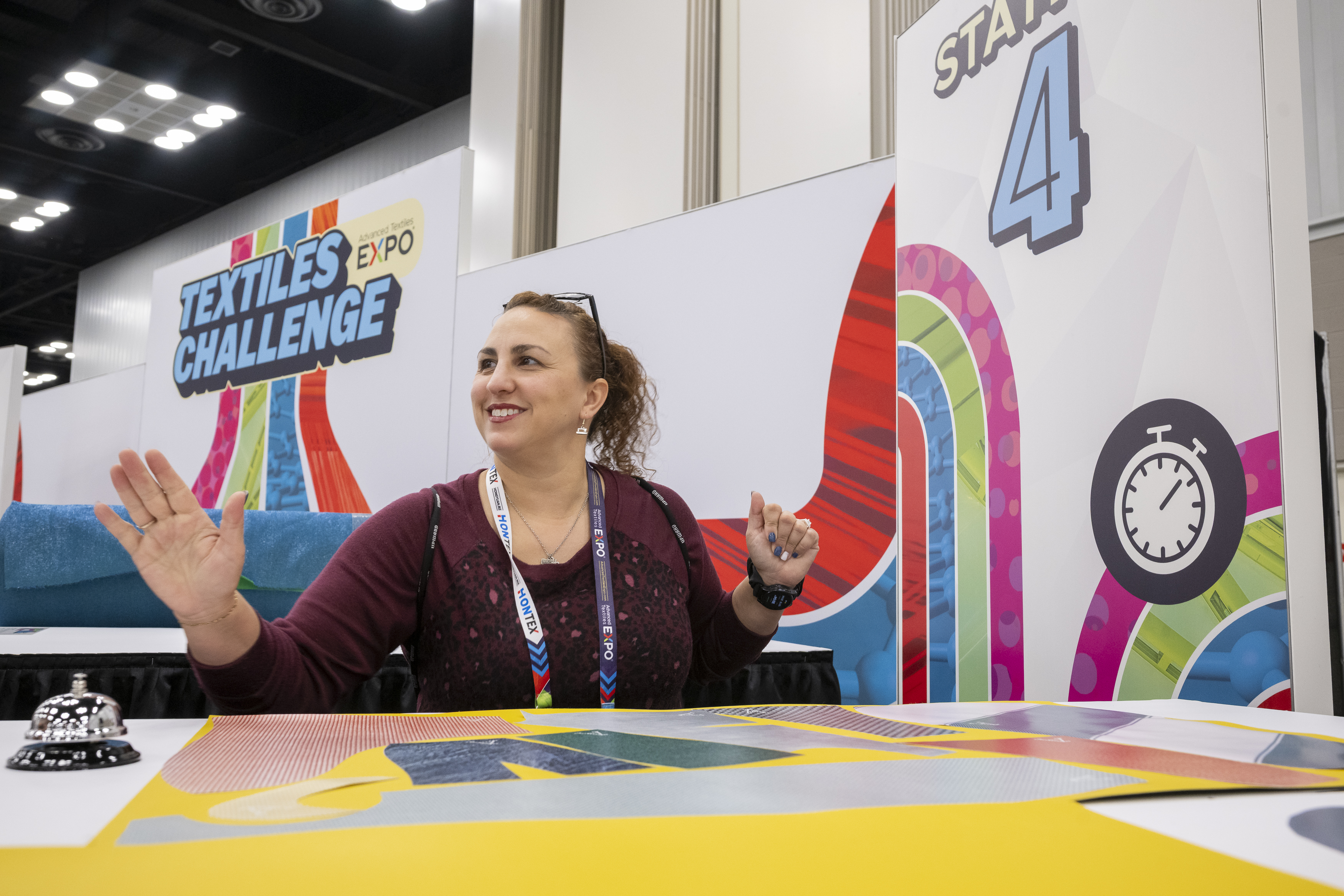 A smiling woman gestures at a colorful booth labeled "Textiles Challenge" during an expo, with vibrant patterns and a timer icon visible.