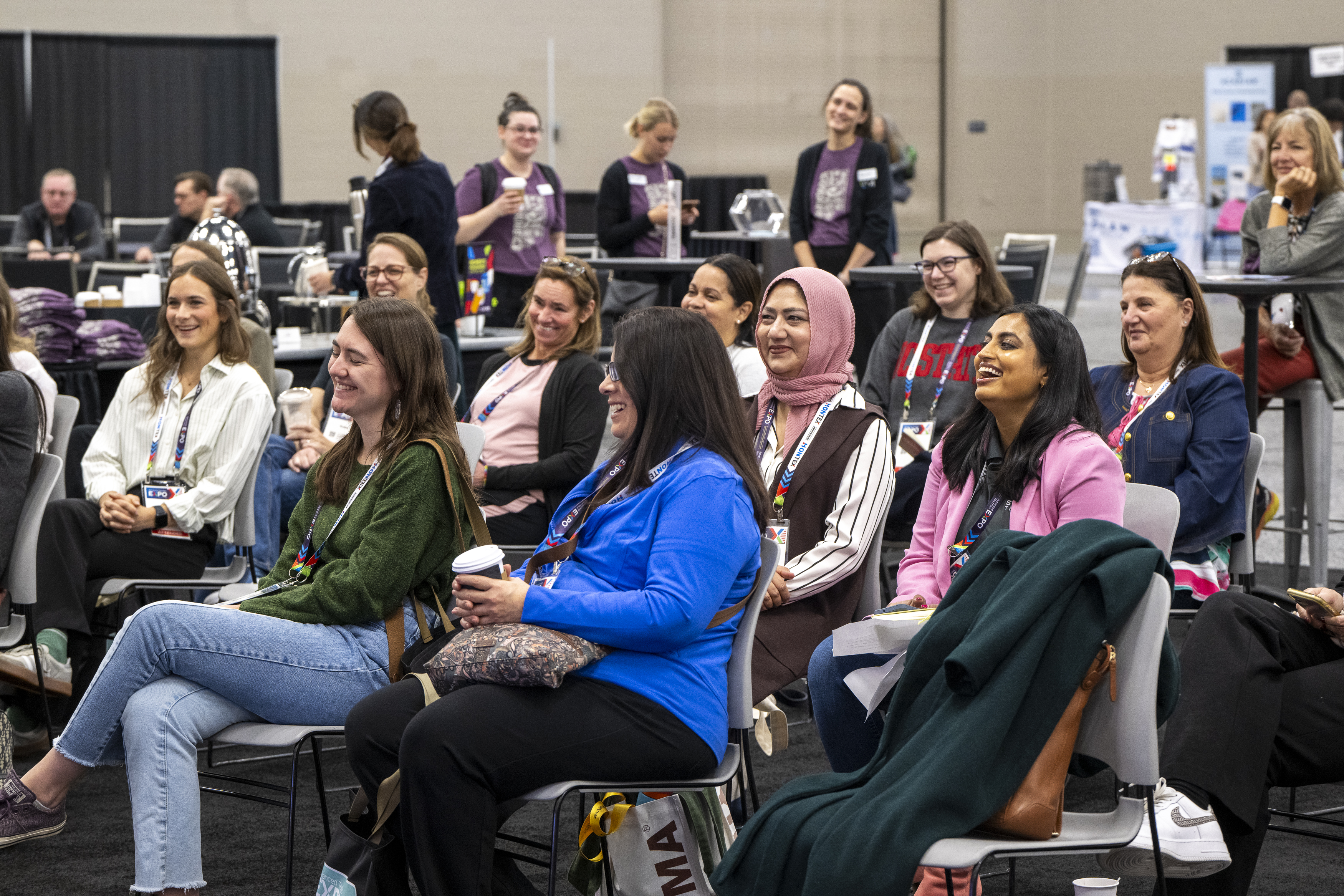 A diverse group of women sit together, smiling and engaged, in a conference setting with black and gray decor and colorful lanyards.