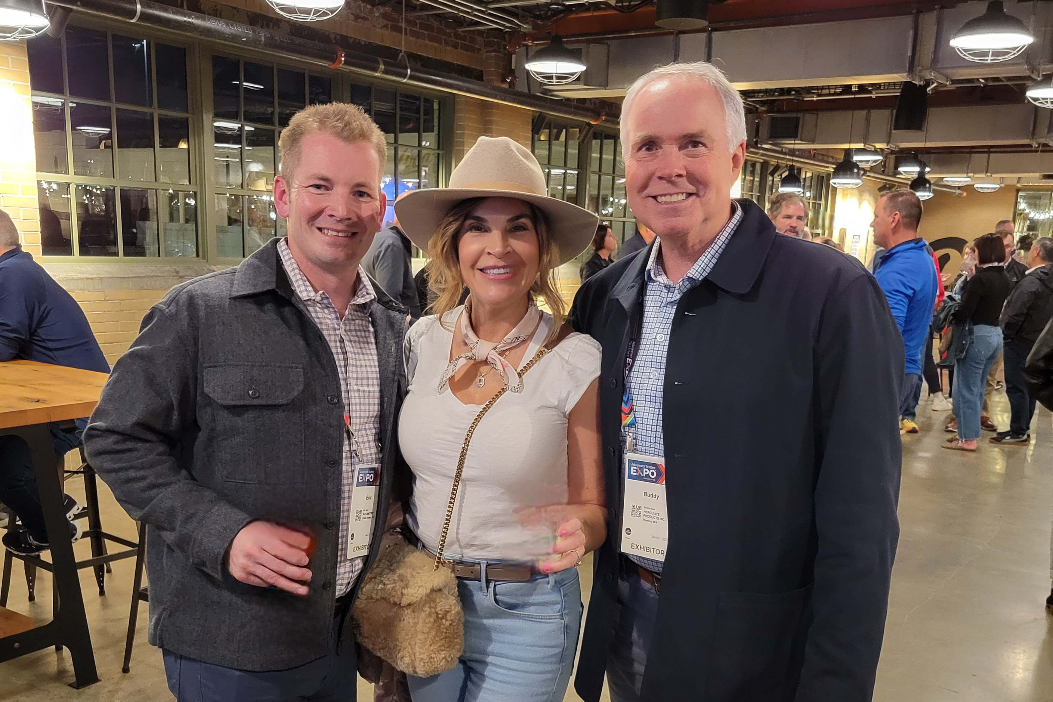 Three individuals pose together in a lively venue, with warm lighting, wearing casual attire. The woman wears a wide-brimmed hat and holds a drink.