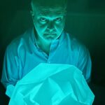 Man with a serious expression holds crumpled light blue fabric, illuminated by a glowing teal light in a dark background.