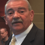 A man with gray hair and a mustache, wearing a suit and tie, looks directly at the camera with a neutral expression.