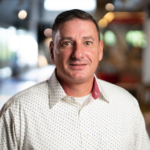 Man with short dark hair and a slight smile, wearing a patterned white shirt with a red collar, set against a blurred background.