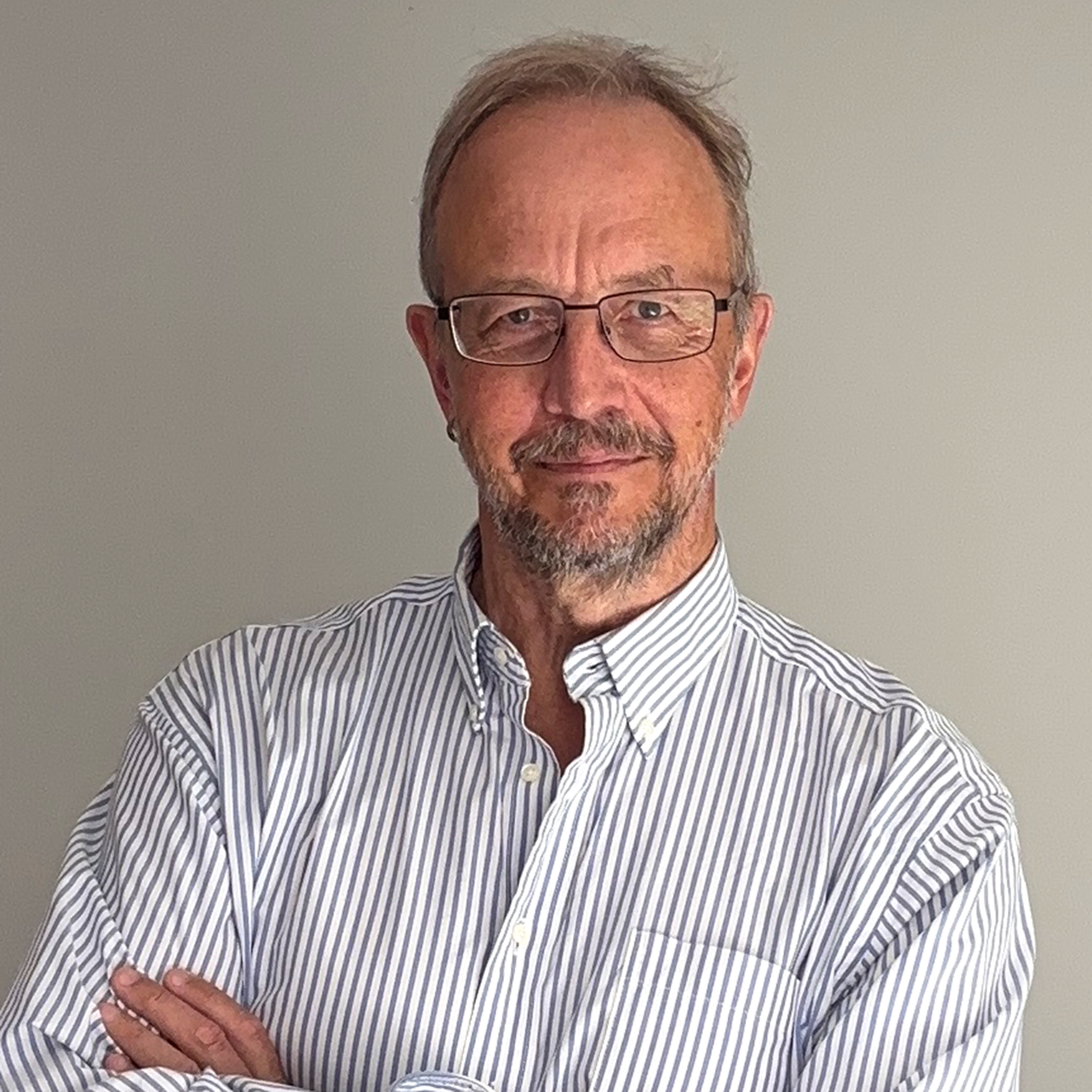 Man with glasses and a light-striped shirt, standing with arms crossed against a neutral background, smiling slightly.