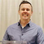 Smiling man in a blue shirt stands behind a clear podium, with a white curtain backdrop, exuding a friendly, approachable demeanor.