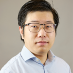 Portrait of a man with short black hair, wearing glasses and a light blue button-up shirt, set against a neutral background.