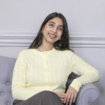 Young woman in a pastel yellow cable-knit sweater smiles while seated on a grey couch, with elegant decor in a light room.
