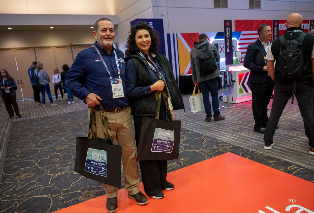 Two attendees smile while standing on an orange event floor, each holding a tote bag; colorful banners and people in the background.