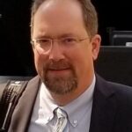 Man with a beard and glasses, wearing a suit and striped tie, smiles confidently against a dark background.