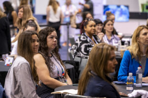 A group of attendees, primarily women with diverse hairstyles and outfits, sit attentively at tables during a conference event.