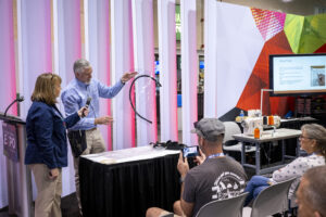 A presenter holds up a clear vinyl piece, demonstrating to an audience at an expo, with colorful backdrop and sewing equipment visible.
