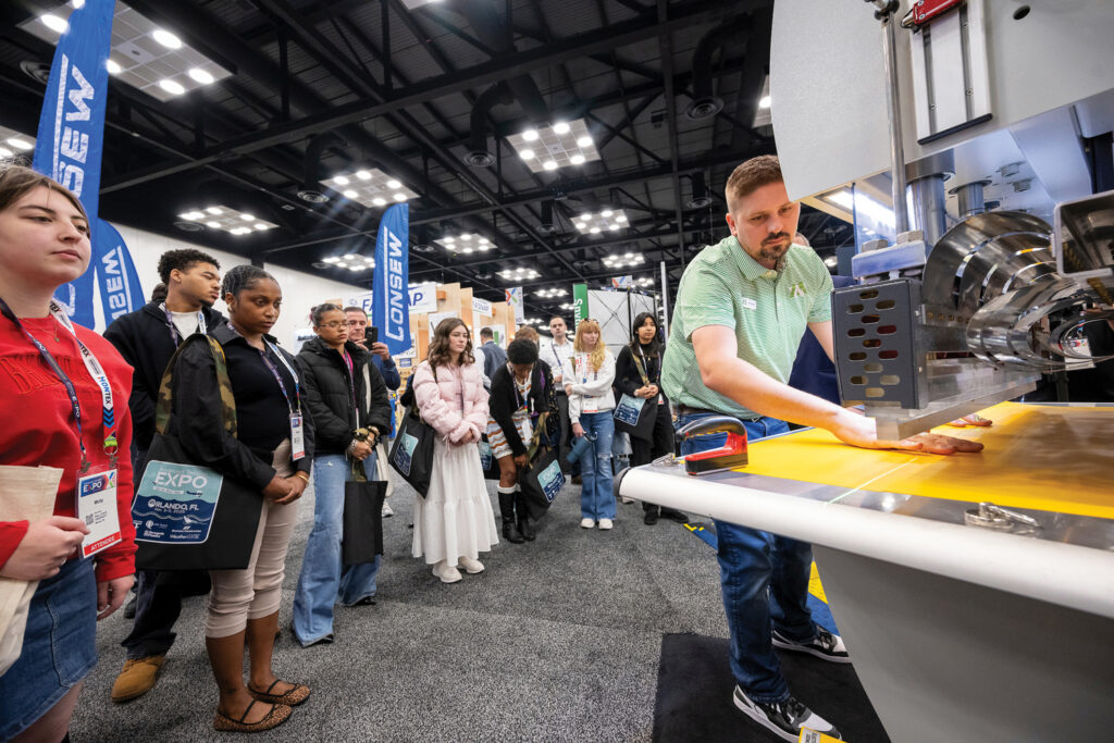 A group of students watches a presenter operate a textile machine, with banners in the background and a focus on the machine's yellow surface.