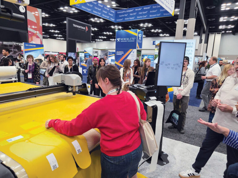 A visitor in a red sweater uses a large yellow fabric printer, surrounded by onlookers and colorful exhibition banners in a busy expo hall.