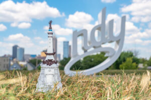 A detailed miniature of the Monument Circle stands in grassy foreground, with a large "indy" sign and a bright blue sky in the background.