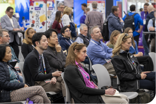 A diverse group of attendees seated and engaged in a conference setting, showcasing a blend of professional attire and colorful badges.