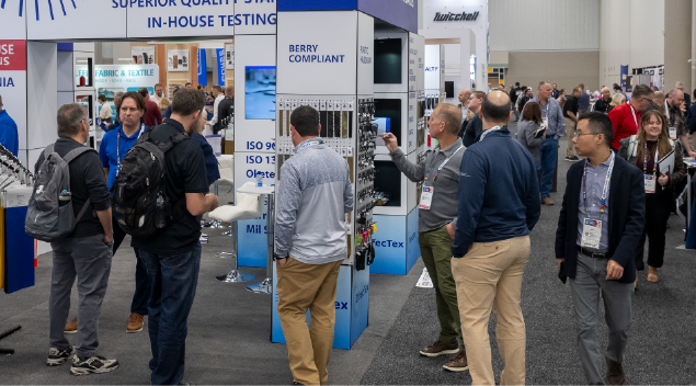 A bustling trade show scene with attendees examining displays, featuring colorful booths and various testing equipment.