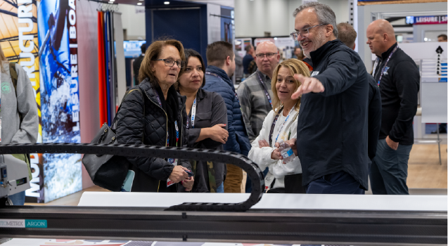 A group of smiling individuals engages with a presenter, discussing a display at an exhibition, with a variety of colorful booths in the background.