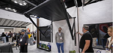 A black retractable awning is showcased in a trade show setting, with attendees engaging nearby and a light-colored backdrop.