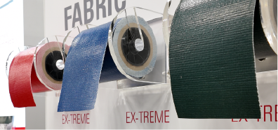 Four rolls of tarpaulin fabric in red, blue, and green hang on display, showcasing their textured surfaces against a light backdrop.