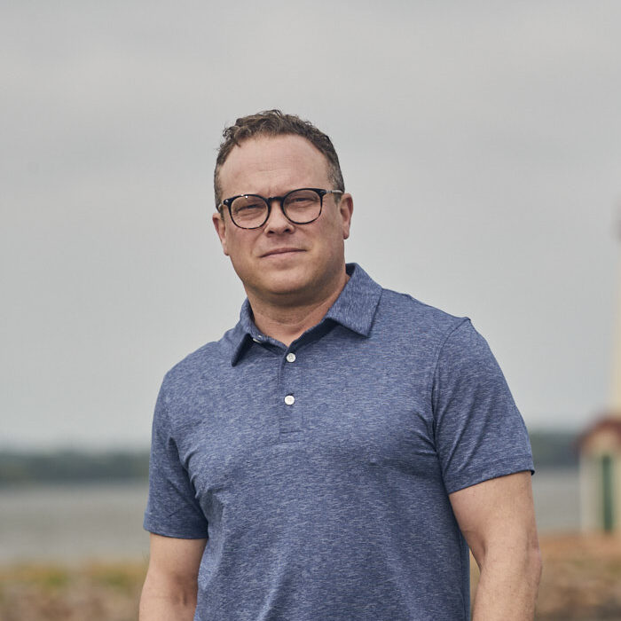 A man in a blue polo shirt stands in front of a lighthouse by the water, with a cloudy sky in the background.