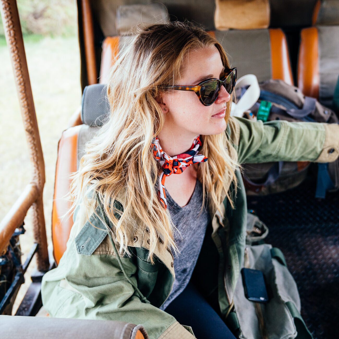 Woman with long blonde hair, wearing sunglasses and a green jacket, sits inside a safari vehicle, looking outside, with a bandana around her neck.