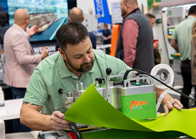 A man in a green striped shirt operates a machine, feeding a vibrant green sheet. Blurred figures and industry booths are in the background.