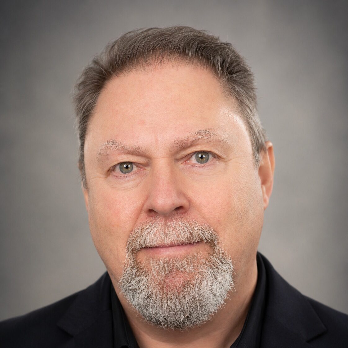 A man with a gray beard and short hair, wearing a black suit jacket over a black shirt, poses against a neutral gray background.