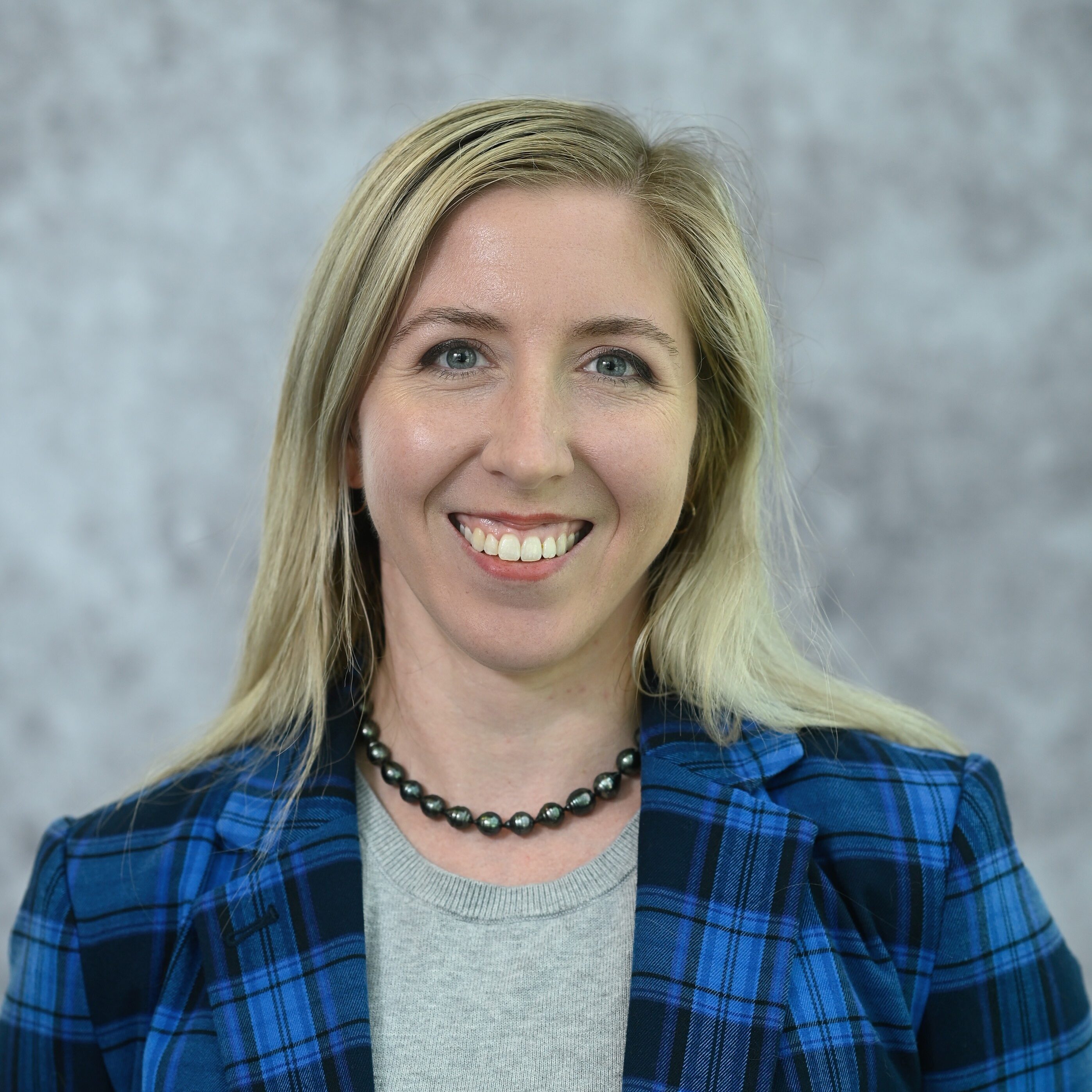 Smiling woman with long blonde hair, wearing a blue and black plaid blazer over a gray sweater, set against a gray backdrop.