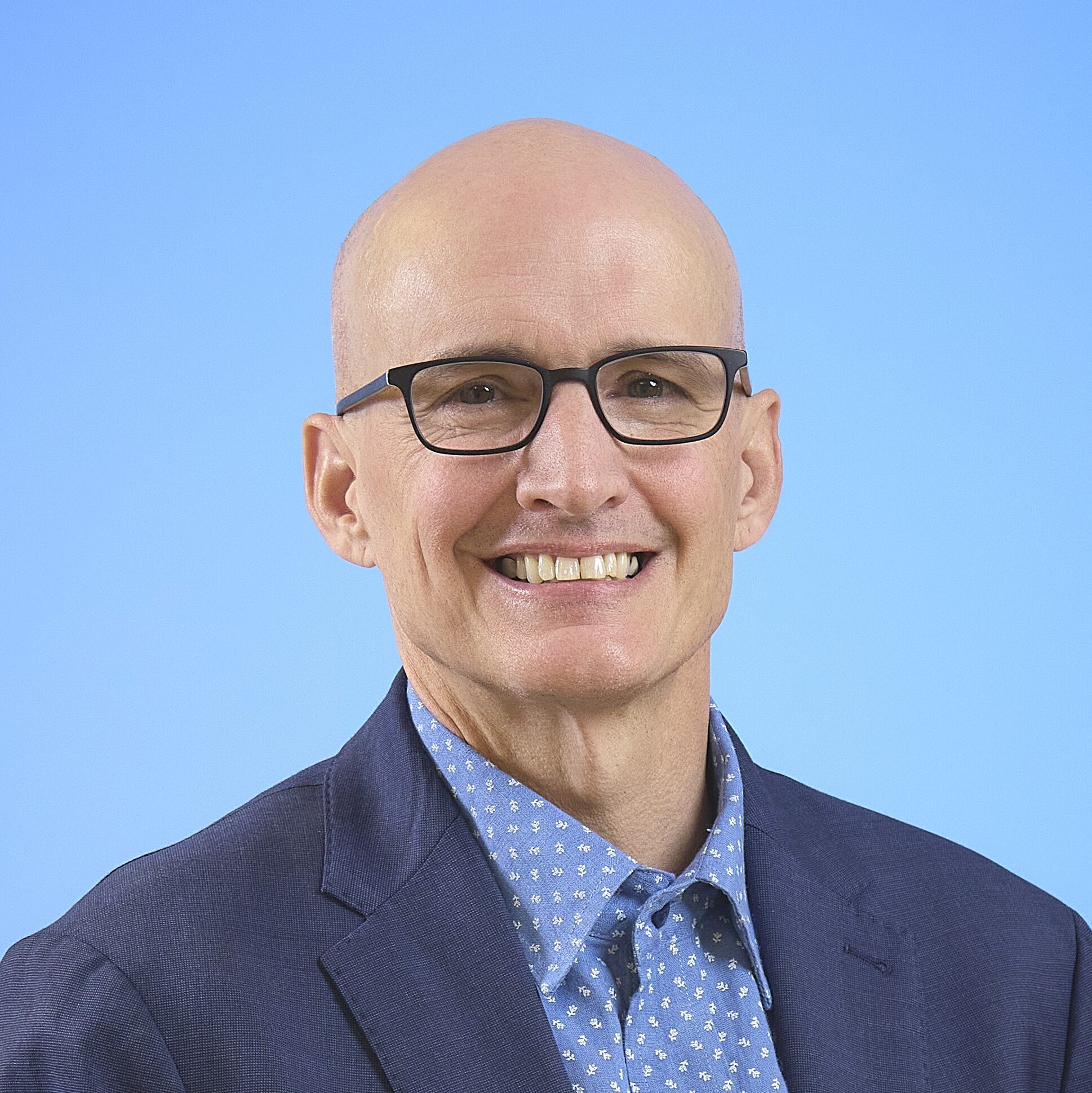 Smiling man with glasses, short bald hair, wearing a blue blazer over a patterned light blue shirt, against a soft blue background.