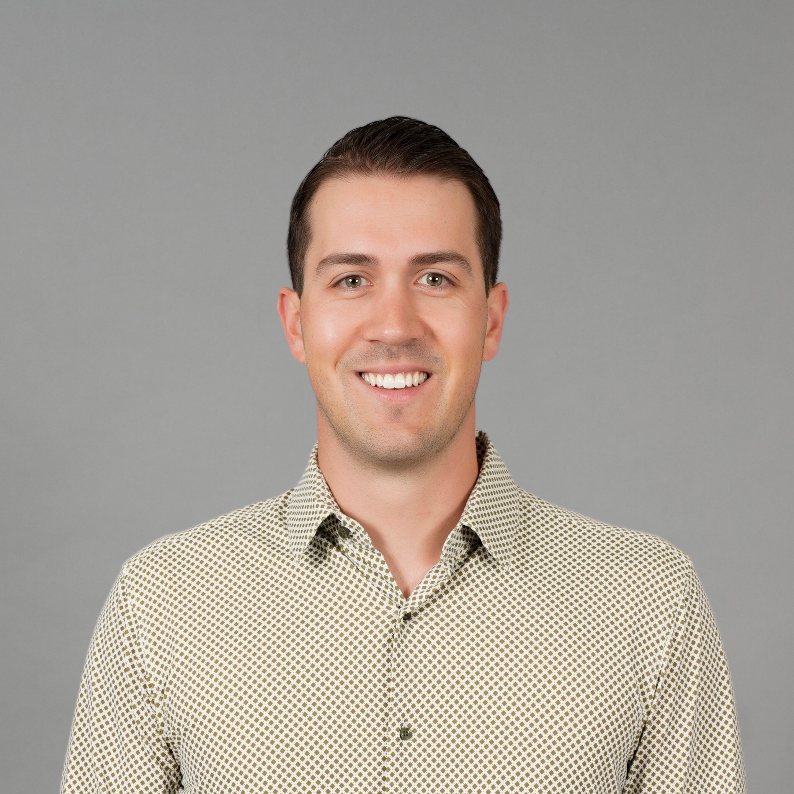 Young man with short hair, smiling, wearing a patterned shirt and jeans, stands with hands in pockets against a gray backdrop.