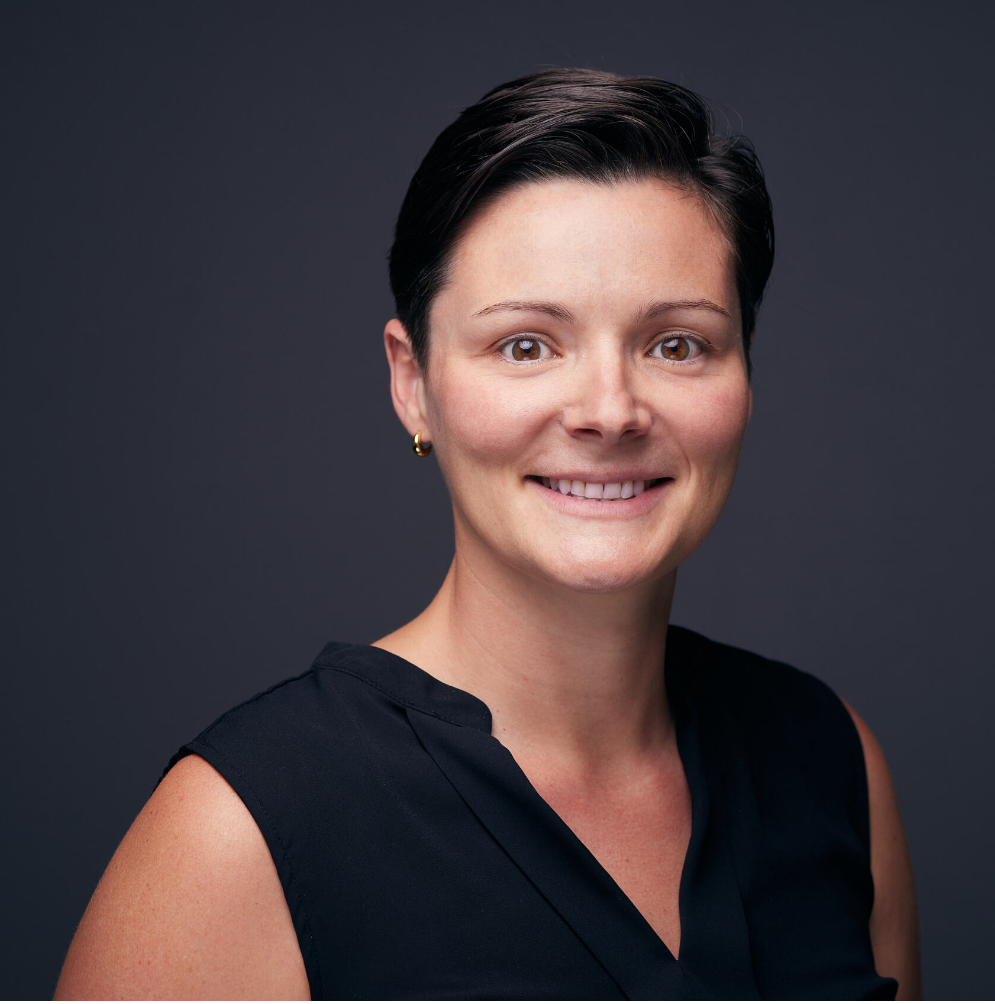 Headshot of a woman with short dark hair, wearing a black top, set against a dark grey background, smiling confidently.