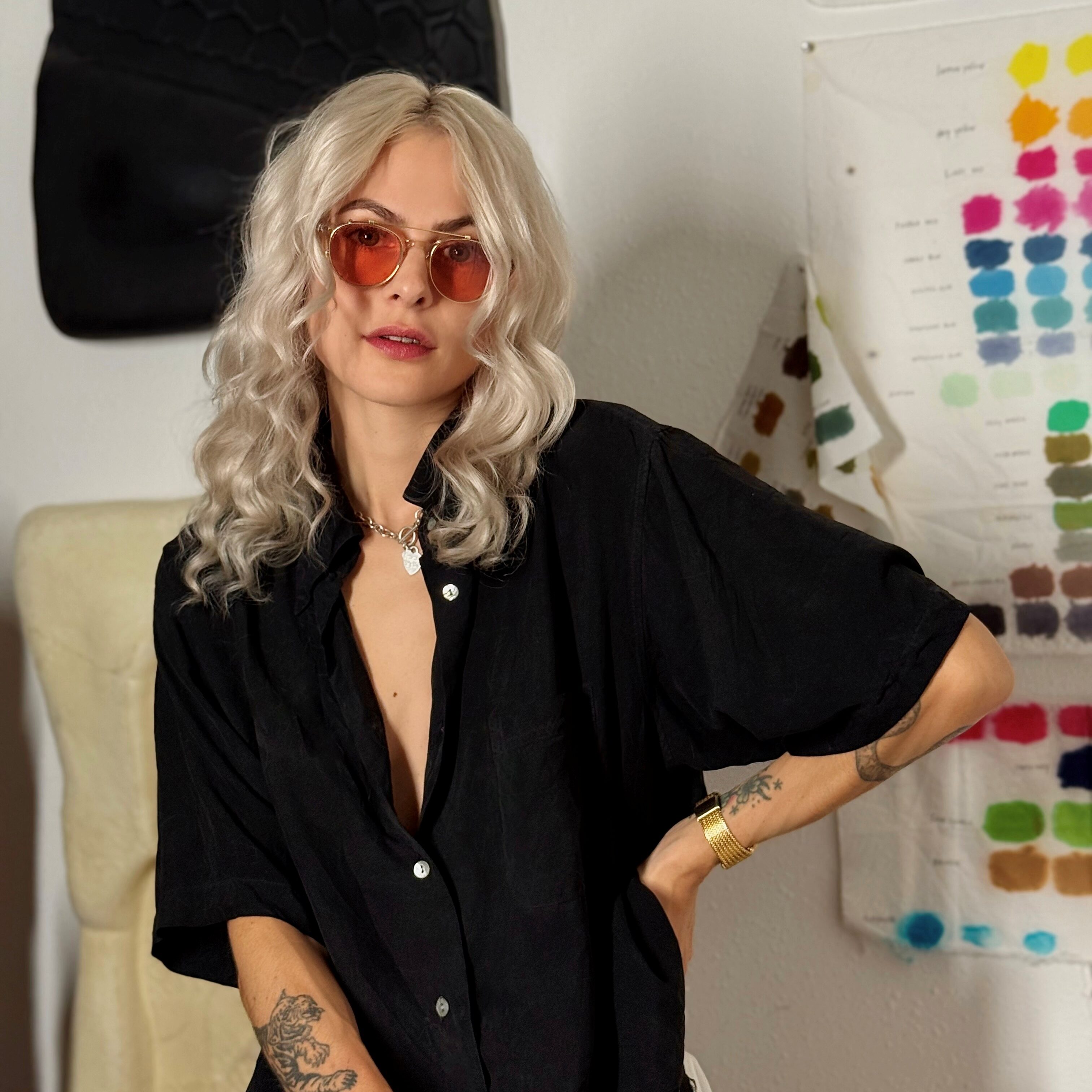 A woman with long, wavy blonde hair wearing orange sunglasses and a black shirt, posing against a colorful backdrop of art swatches.