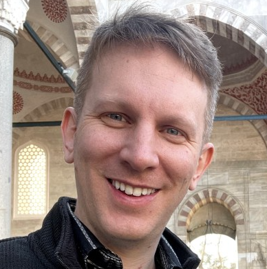 Smiling man with short, light hair, wearing a black jacket, stands in a mosque with intricate architectural details in the background.