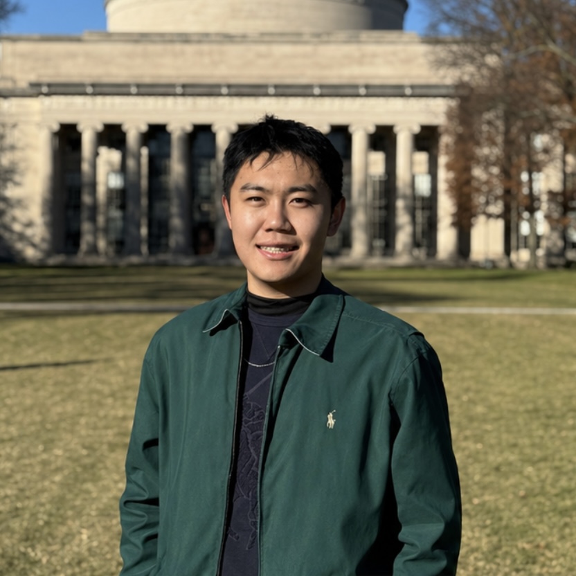 A young man in a green jacket stands on grass in front of a large, classical building with columns and wide steps.