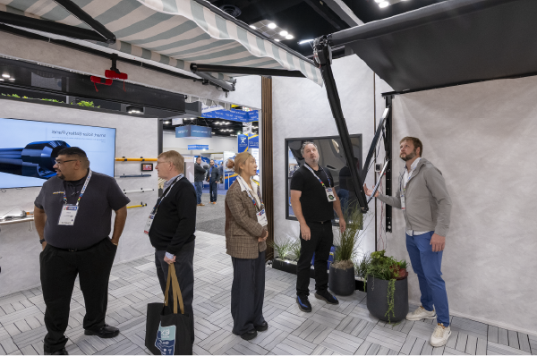 A group of four people interacts under a retractable striped awning in a modern booth setting, with plants and a display screen in the background.