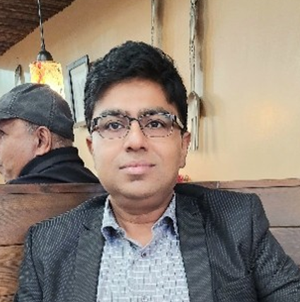 A man with glasses and a dark blazer sits at a restaurant table, with warm lighting and a blurred figure in the background.